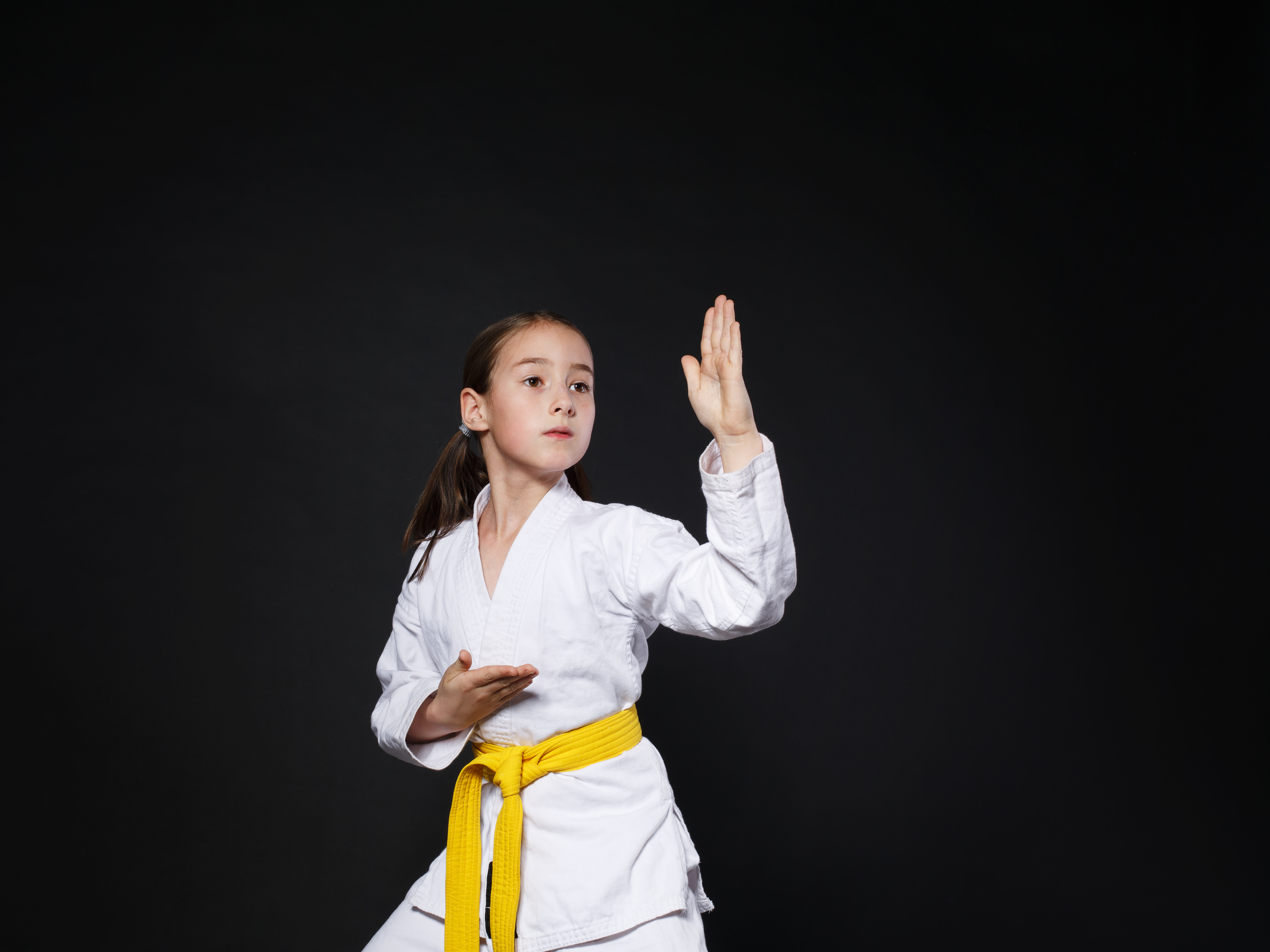 Little girl in karate suit kimono in studio at black background. Female child shows judo or karate stans in white uniform with yellow belt. Individual martial art sport for kids. Waist up portrait